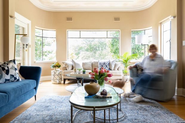 Bright, warmly styled living room with curved bay-window glazing, a white sofa, blue armchair, round glass coffee tables, layered rugs, indoor plants, and an ornate recessed ceiling detail. The room has pale timber flooring, warm beige walls, and abundant natural light.