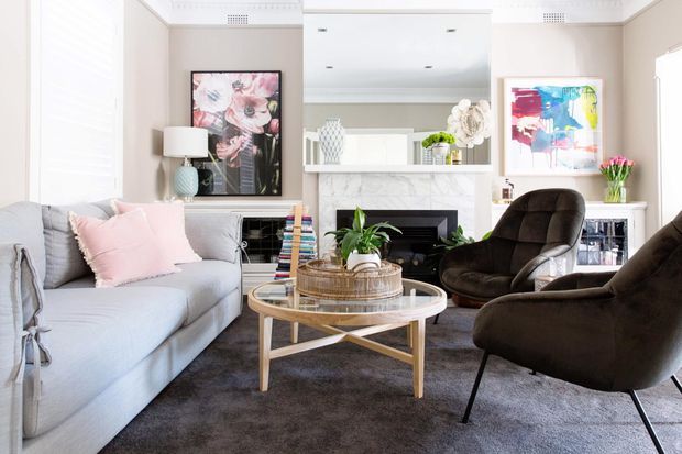 Bright, refined living room with a pale upholstered sofa, blush cushions, dark sculptural lounge chairs, a round timber-and-glass coffee table, fireplace, mirrored mantel, floral artwork, and soft neutral finishes. The room combines classic architectural detailing with contemporary furnishings and colourful accents.