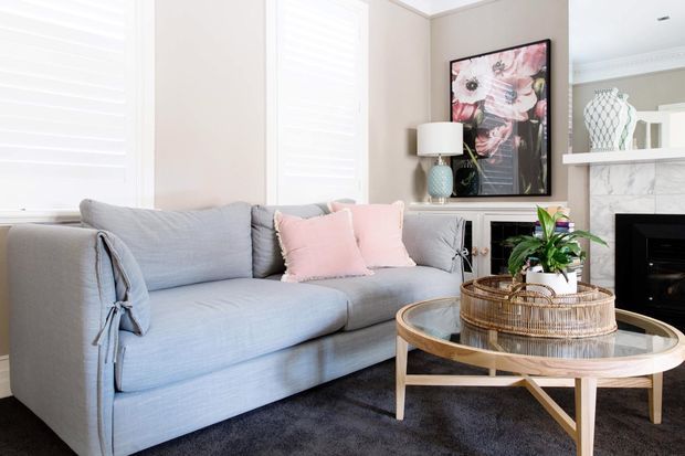 Bright, refined living room with a light blue-grey upholstered sofa, blush cushions, dark rug, and a round glass-topped timber coffee table. The room features plantation shutters, pale neutral walls, a marble-look fireplace surround with white mantel shelving, a framed floral artwork, and a decorative indoor plant.