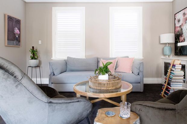 A bright, compact living room with a grey sofa and matching upholstered lounge chairs arranged around a round timber-and-glass coffee table. Pale neutral walls, white plantation shutters, dark carpet, soft pastel cushions, indoor plants, a floor lamp, stacked books, and wall artwork create a comfortable, layered setting.