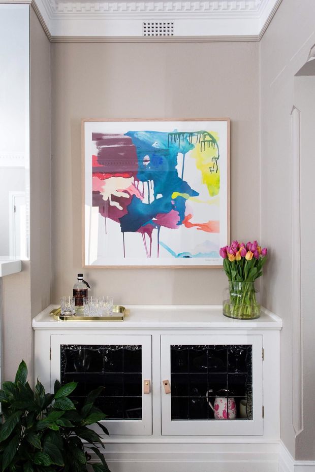 A refined interior vignette featuring a white built-in cabinet or sideboard with black glossy tiled inset doors, a pale stone or painted benchtop, and brass-toned tray hardware. Above it hangs a colourful abstract artwork, while a vase of tulips and a leafy indoor plant add organic accents. The room has light taupe walls, white architectural trim, ornate geometric cornice detailing, and a bright traditional character.