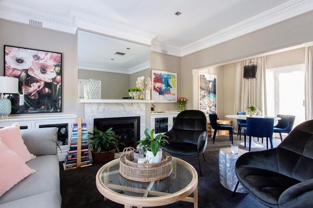 Bright, elegant living room with a light grey sofa, black upholstered lounge chairs, round glass coffee table, fireplace with white mantel, built-in cabinetry, large mirror, decorative artwork, and a dining area visible beyond. The room features ornate ceiling cornices and trims, neutral taupe walls, and layered contemporary furnishings in a refined classic setting.