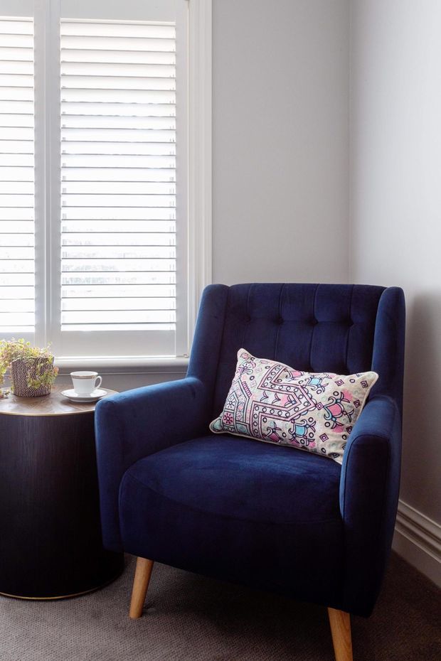 A bright, compact reading nook featuring a deep navy-blue upholstered wingback armchair with button tufting and tapered timber legs. A patterned decorative cushion rests on the chair, beside a small dark round side table holding a potted plant and coffee cup. White plantation shutters cover the window, with pale walls, white trim, and neutral carpet.