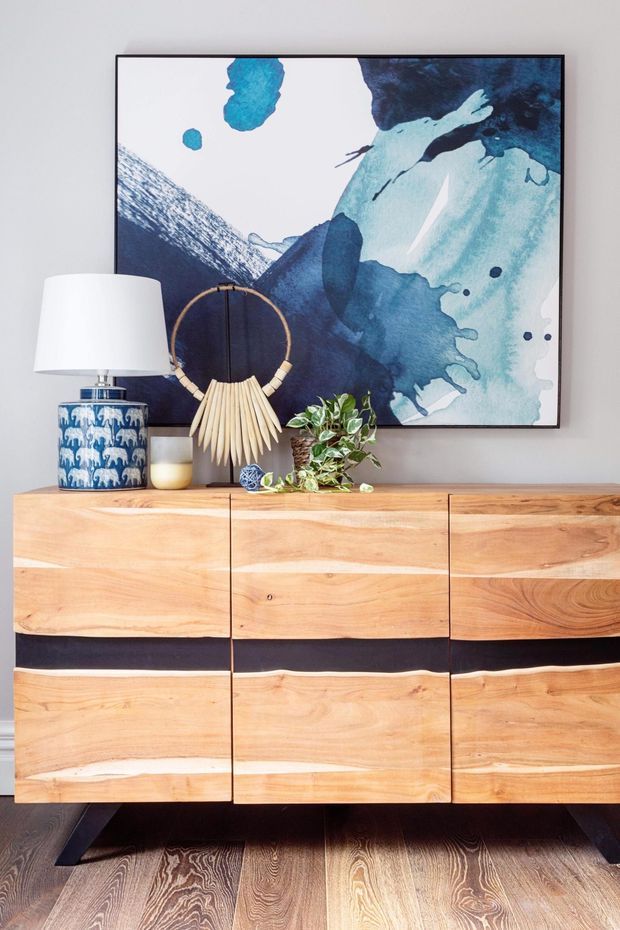 A styled interior vignette featuring a mid-century-inspired wooden sideboard with natural timber grain and dark recessed drawer bands, set against a light wall. A blue and turquoise abstract artwork hangs above it, accompanied by a white table lamp with a blue patterned ceramic base, a sculptural woven loop lamp, a small plant, and decorative objects. Dark timber flooring is visible below.