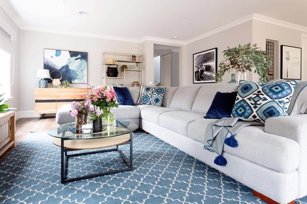 Bright, spacious living room with a large light-grey sectional sofa, blue patterned rug, glass coffee table, blue and white cushions, floral arrangement, timber sideboard, open shelving, framed artwork and indoor greenery. The room has pale walls, white crown moulding and abundant natural light.