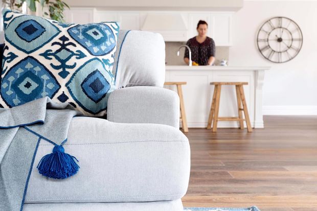 Bright open-plan living area with a pale upholstered sofa in the foreground, accented by bold blue-and-white geometric cushions and a matching throw with tassel detail. A white kitchen with a peninsula, stools, cabinetry, and a person preparing food is visible behind it. The room features warm timber flooring, white walls and trim, and a large round wall clock.