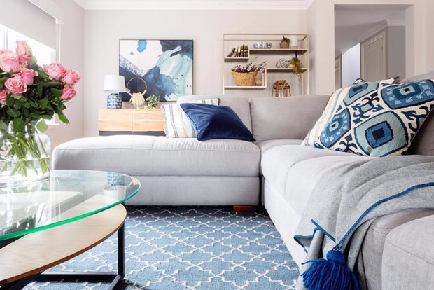 Bright, inviting living room featuring a large light-grey sectional sofa, blue patterned cushions and throw, a blue-and-white geometric rug, a glass-topped timber coffee table, open shelving, artwork, and a floral arrangement. The room has pale walls, white trim, and a contemporary transitional feel.