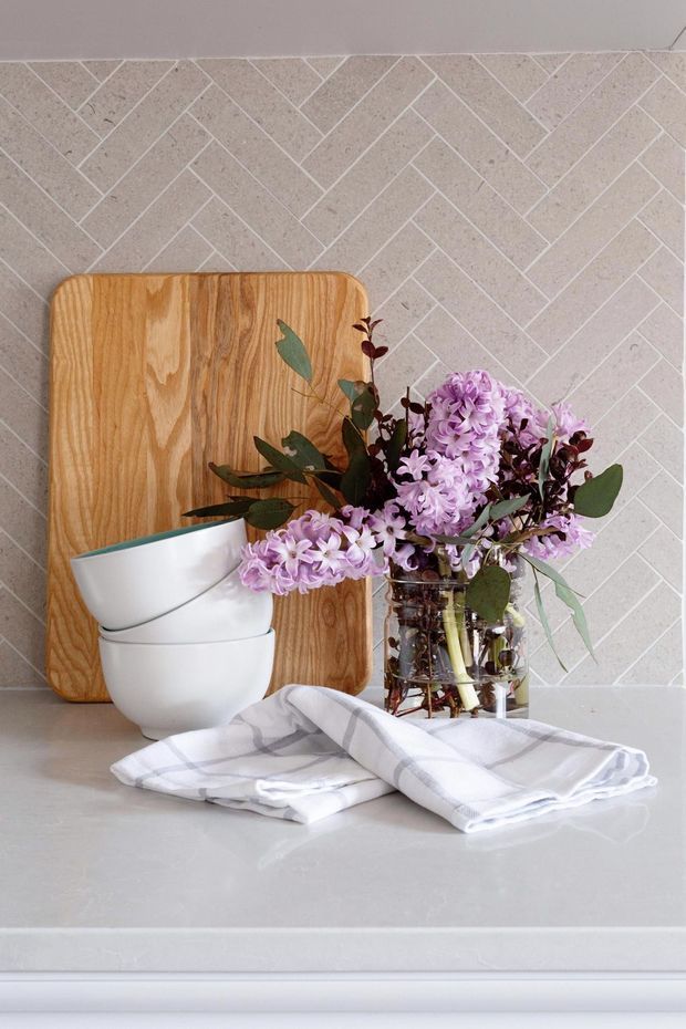 A styled kitchen or dining benchtop vignette featuring a pale stone-look countertop, white ceramic bowls, a wooden chopping board, folded white-and-grey tea towels, and a glass vase of purple flowers. The backsplash has a light beige geometric herringbone tile pattern, creating a soft, refined still-life composition.