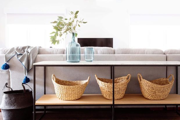 Bright, airy living room with a pale upholstered sectional sofa, a slim dark-framed console or open shelving unit, woven baskets, and glass vases with leafy branches. White walls and window treatments create a light neutral backdrop, with natural woven textures adding warmth.
