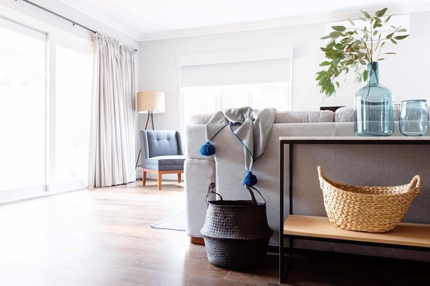 Bright, airy living room with a large neutral sectional sofa, pale timber flooring, floor-to-ceiling glazing with sheer curtains, and a slim dark-framed console displaying blue glass vessels and a woven basket. A blue accent armchair and floor lamp sit near the window.