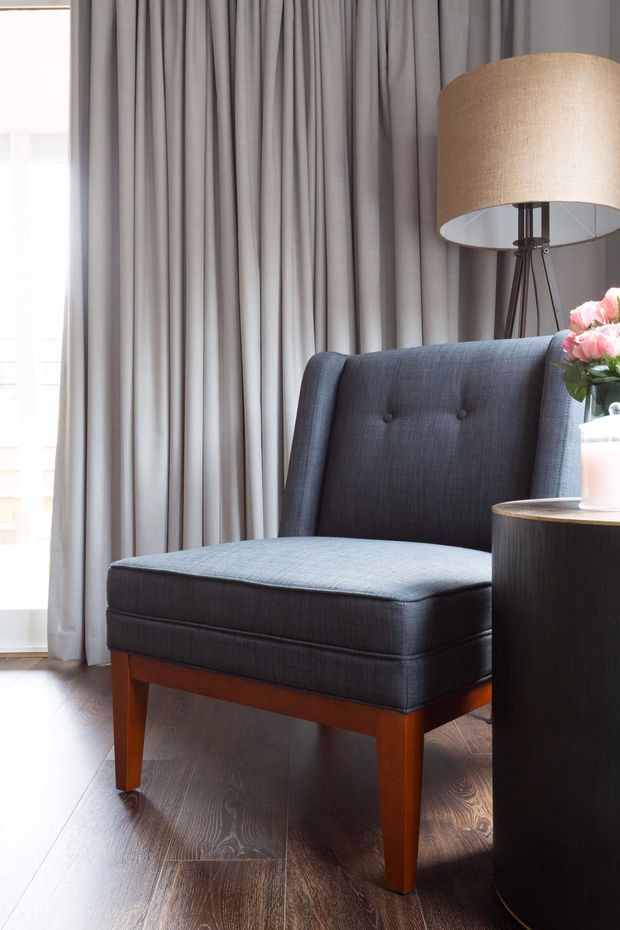 A softly lit interior vignette featuring a tailored charcoal-blue upholstered accent chair with button detailing and warm wooden legs, positioned beside a dark wood round side table. Floor-to-ceiling grey curtains, a large fabric-shaded floor lamp, flowers, and dark timber flooring create a refined, contemporary setting.