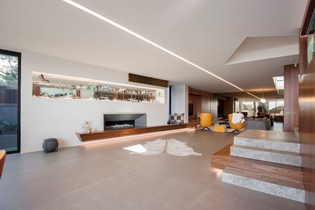 A spacious contemporary open-plan living area with a polished concrete floor, a linear built-in fireplace and display niche, warm timber wall panelling and cabinetry, and broad glazing. A sculptural timber staircase with stone-tread steps rises at the right, while lounge and dining areas extend toward the rear. A slim linear ceiling light and recessed ceiling detailing reinforce the minimalist architectural design.