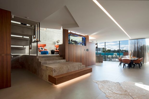 A spacious contemporary open-plan interior with polished concrete flooring, a sculptural stepped stair and timber-clad feature wall. The living and dining areas open onto full-height glazing and a balcony with water views. Exposed concrete, dark timber joinery, linear recessed lighting, colourful lounge chairs, and a large sculptural rug create a refined modern atmosphere.