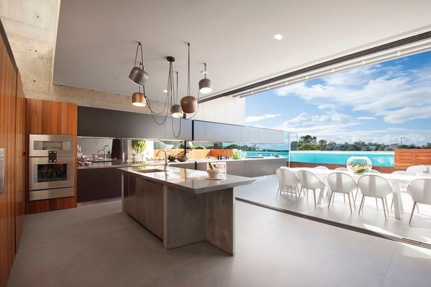 Contemporary open-plan kitchen and dining space with a large central island, dark and timber cabinetry, integrated stainless-steel appliances, pendant lighting, and extensive sliding glazing opening to a bright terrace with water views. The interior uses polished concrete surfaces and a restrained neutral palette.