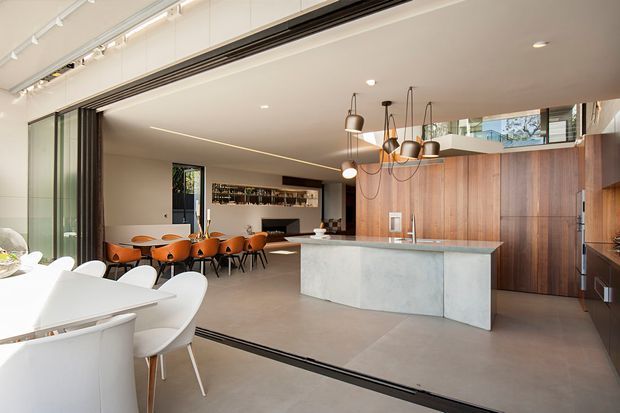 A contemporary open-plan kitchen and dining area with a large pale stone island, dark timber cabinetry, integrated appliances, and a row of sculptural pendant lights. The adjoining dining space includes a long table with orange upholstered chairs, while extensive sliding glass doors open the room toward an outdoor area. Track lighting, recessed ceiling lights, polished concrete-style flooring, and warm timber wall finishes create a refined indoor-outdoor setting.
