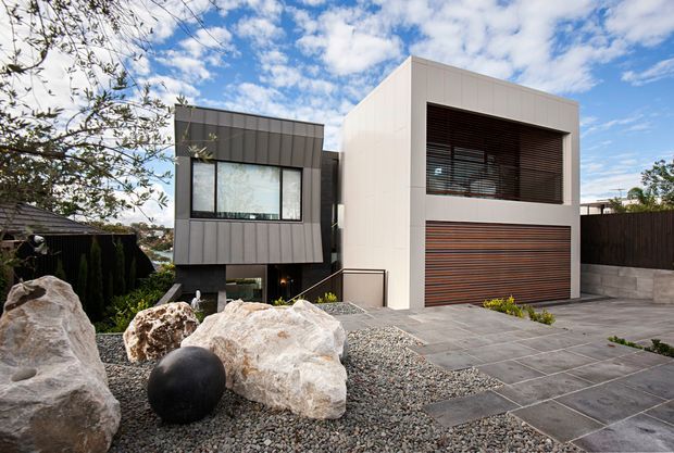 Contemporary two-storey residential facade with a crisp white rendered upper volume, dark grey vertically clad projecting section, large horizontal windows with dark timber-toned louvres, and a matching timber-toned garage door. A paved driveway and minimalist gravel-and-boulder landscape occupy the foreground.