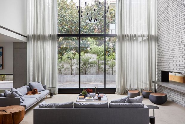 A spacious contemporary living room with a double-height glazed wall framed in black metal and dressed with sheer white curtains. Multiple grey sofas surround a low coffee table, while a white-painted brick feature wall contains a long fireplace ledge and television. Light flooring, restrained furnishings, and abundant greenery visible beyond the glazing create an airy, modern atmosphere.
