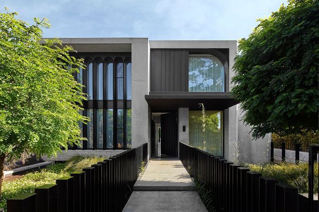 Front exterior view of a contemporary two-storey residence, with a strong geometric facade, dark vertical metal fencing, a raised pedestrian approach, large arched-top glazing, and textured grey rendered walls. Mature greenery frames the entry.