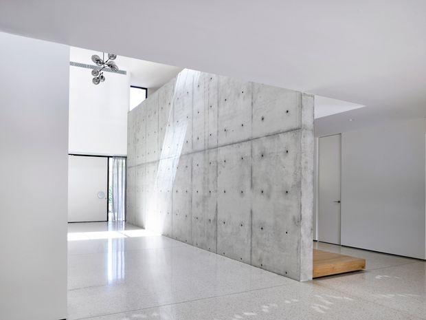A bright, minimalist interior with a large exposed concrete feature wall dividing an open-plan space. The polished light floor reflects daylight from tall glazing, while a warm timber platform or stair element emerges behind the concrete wall. White walls, clean lines, and a sculptural modern light fixture reinforce the gallery-like architectural character.