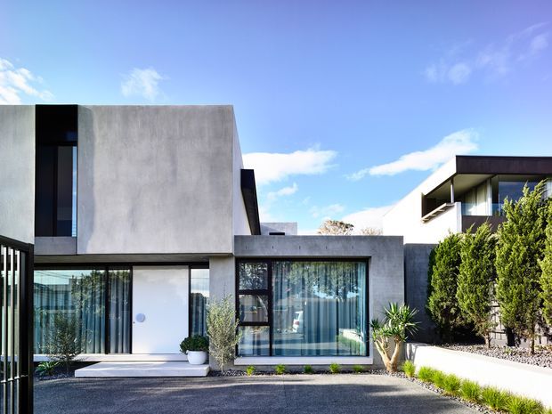 Exterior view of a contemporary residential house with strong geometric forms, smooth grey concrete or rendered walls, extensive floor-to-ceiling glazing, dark window framing, and a flat roofline. The landscaped frontage includes clipped shrubs, gravel, and a paved driveway under a bright blue sky.