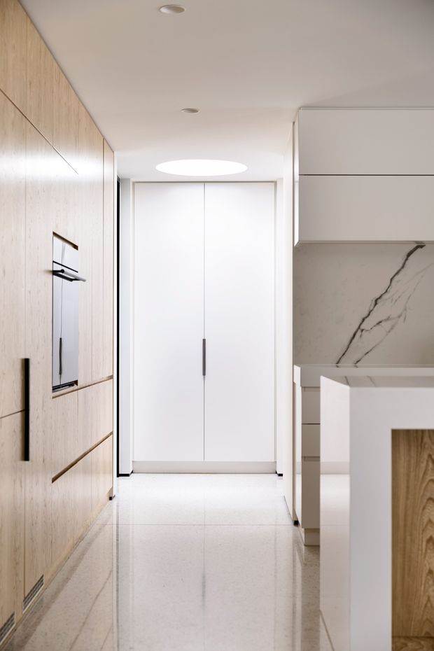 A bright, narrow contemporary kitchen corridor with full-height light timber cabinetry on the left, integrated appliances, white handleless cupboards at the end, and a white island or benchtop on the right. A large marble-look splashback with bold dark veining provides the main feature, while polished pale flooring and recessed ceiling lights create a clean, refined appearance.
