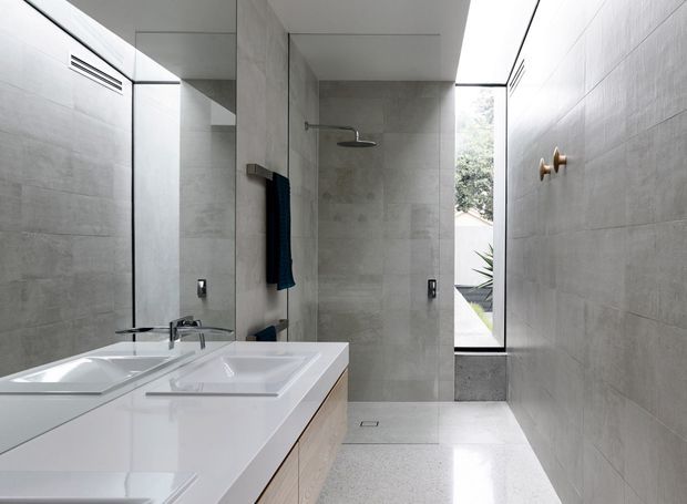 Contemporary minimalist bathroom with a long white double vanity, integrated basins and chrome tapware, pale grey tiled walls and floor, a frameless glass shower area, and a large mirror. A narrow full-height window brings in natural light and views of greenery, while subtle brass wall hooks provide a small accent.