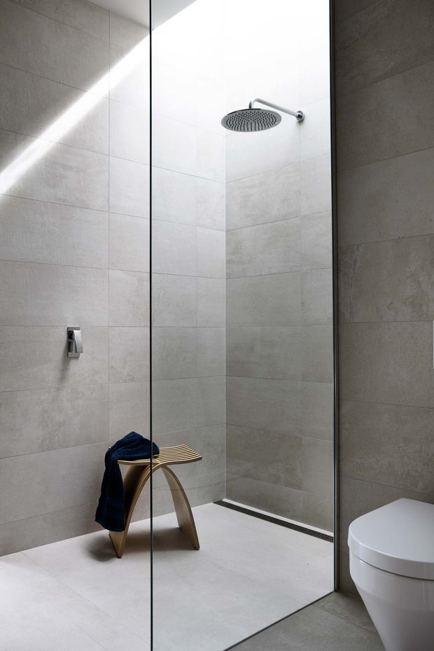 A minimalist contemporary bathroom featuring a large walk-in glass shower enclosure, pale grey stone-look wall and floor tiles, a round rainfall showerhead, linear floor drain, and a white toilet partially visible at the right. A small curved wooden stool with a dark towel adds warmth to the otherwise clean, neutral space.