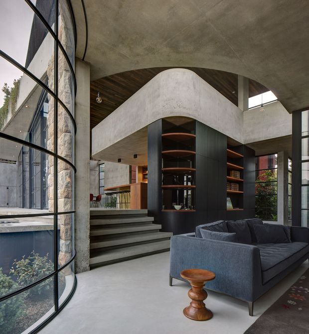 A dramatic contemporary interior with a sunken living area, sculptural curved concrete mezzanine or stair enclosure, exposed concrete ceiling and floor, dark built-in shelving, and warm timber ceiling accents. A large curved glazed wall brings in abundant natural light, while a deep blue-grey sectional sofa and small turned timber side table furnish the foreground. Steps lead upward toward a compact kitchen or bar area.