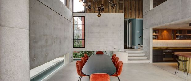 A dramatic open-plan contemporary dining and living space with a long dark dining table surrounded by sculptural burnt-orange leather chairs. Exposed concrete walls and floors create a gallery-like shell, while warm timber wall cladding and integrated illuminated shelving form a bar or kitchen zone at the rear. A tall glazed opening, split levels, and a sculptural clustered pendant contribute to the spacious, high-volume interior.