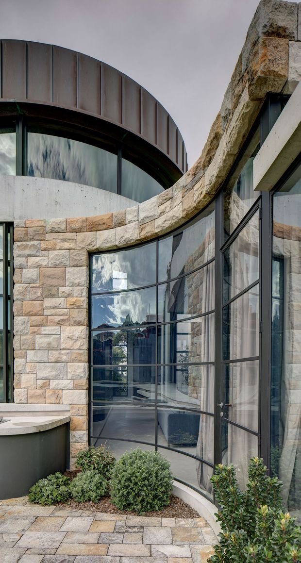 Exterior architectural detail of a sculptural building with curved stone masonry walls, expansive black-framed glazed windows and doors, a curved metal roof or parapet element, and a paved courtyard with low planted landscaping. The design combines heavy natural stone, concrete, dark metal framing, and reflective glazing in a contemporary composition.