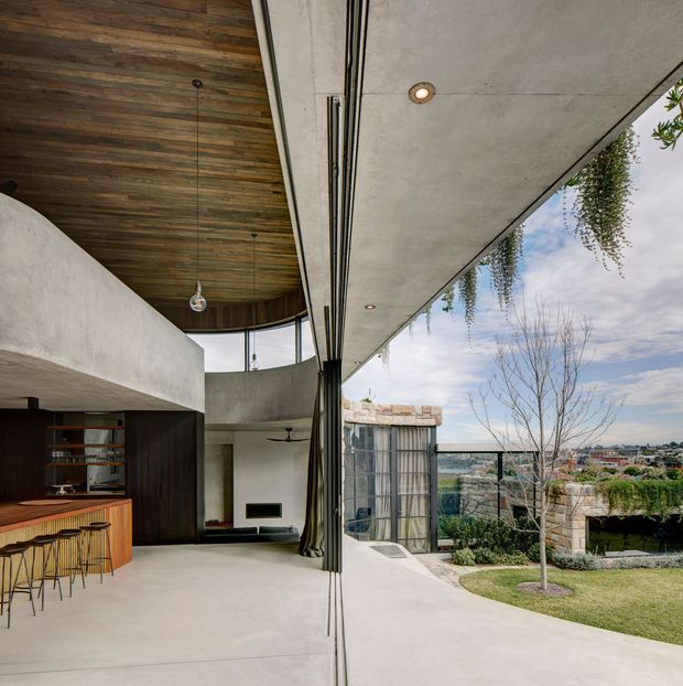 A dramatic contemporary open-plan interior with a built-in dark timber bar and shelving, concrete floors and walls, exposed timber ceiling, and a large sliding glass opening to a landscaped garden and distant urban view. The tall glazed opening, suspended pendant light, recessed downlights, and curved upper-level form create a strong indoor-outdoor connection and an expansive, sculptural atmosphere.