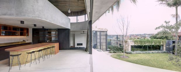 A sophisticated indoor-outdoor entertaining space with a built-in bar and kitchen opening onto a covered terrace and landscaped garden. The interior features a long curved timber-toned counter with bar stools, dark fixed cabinetry, concrete walls and ceiling planes, and expansive glazing or openable panels connecting to the exterior. The covered terrace has a timber-lined ceiling, while the garden includes mature planting, lawn and a hedge-covered structure. The overall composition is clean-lined, spacious and contemporary.
