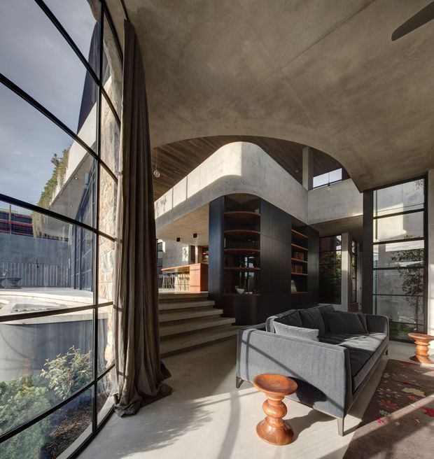 A dramatic double-height contemporary living room with exposed concrete ceilings, sculptural curved concrete openings, dark built-in shelving, and large black-framed floor-to-ceiling glazing. A grey sofa faces into the room, while timber cabinetry and shelving warm the otherwise concrete and charcoal palette. Steps lead to an elevated kitchen or dining area, and strong sunlight creates defined shadows across the polished floor.