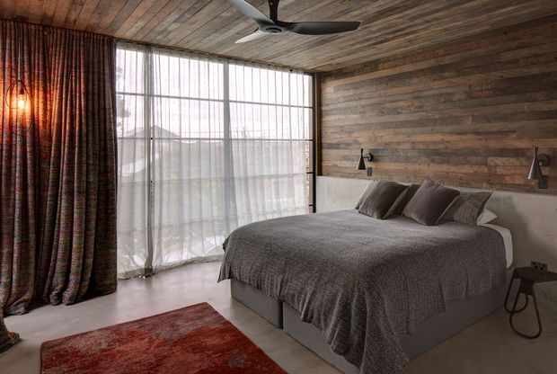 A warm, rustic bedroom with a low-profile upholstered bed dressed in charcoal-grey bedding, set against a reclaimed timber wall and matching timber ceiling. Floor-to-ceiling glazing with sheer curtains provides abundant filtered daylight, complemented by dark patterned drapes, wall-mounted bedside reading lamps, a black ceiling fan, and a red area rug.