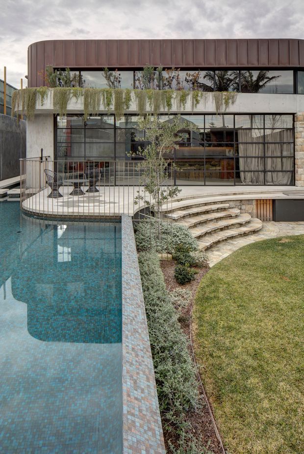 Exterior view of a contemporary multi-level residence centered on a landscaped swimming pool and curved outdoor terrace. The building features extensive floor-to-ceiling glazing with dark metal frames, a rounded upper level clad in dark metal panels, a pale concrete balcony planter, stone retaining and stair elements, and cascading greenery. A curved paved deck with outdoor dining furniture overlooks the pool, with lawn and planted garden beds in the foreground.