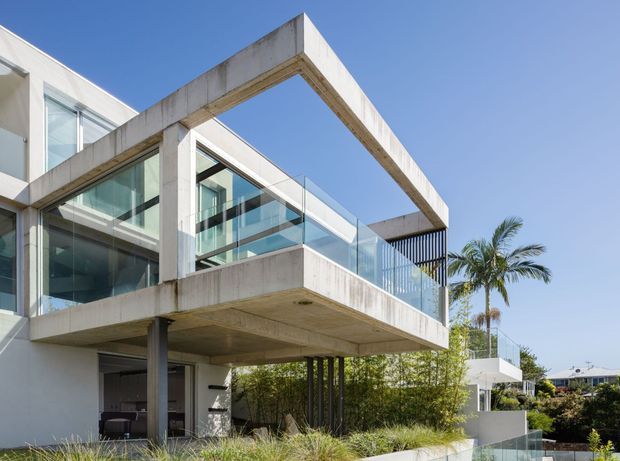 Exterior view of a contemporary multi-level residence with projecting concrete-framed balconies, extensive floor-to-ceiling glazing, transparent glass balustrades, and dark vertical screening. Tropical palms and landscaped planting surround the structure under a clear blue sky.