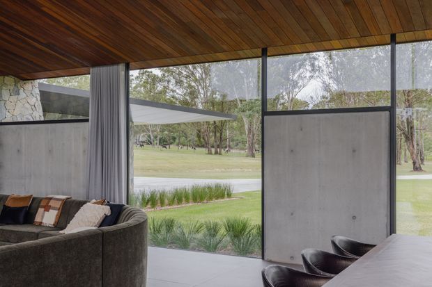 A contemporary open-plan interior with floor-to-ceiling glazing overlooking a landscaped park-like setting. The room features a dark timber-lined ceiling, polished concrete-look floor, exposed concrete wall panels, slim dark window frames, a large curved dark sofa, and a dining table with black leather chairs. Full-height curtains frame the glazing, creating a strong indoor–outdoor connection.
