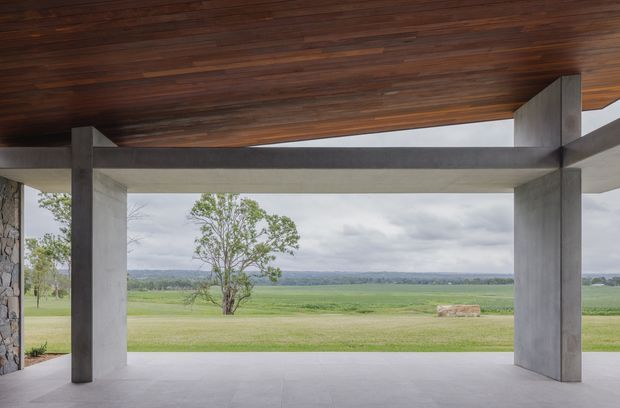 A minimalist covered outdoor pavilion or terrace framed by large exposed concrete columns and beams, with a warm dark timber soffit overhead. The open-sided structure overlooks expansive green rural fields and a distant tree-lined horizon. A stone-clad wall is partially visible at the left edge, while the broad pale floor and restrained material palette create a strong indoor-outdoor architectural connection.
