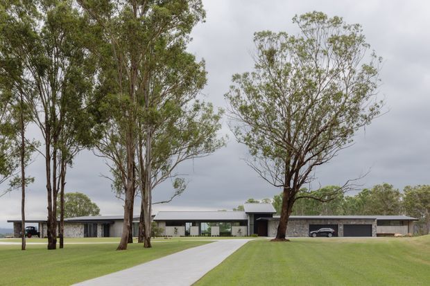A wide exterior view of a long, low-slung contemporary building set in a broad manicured lawn. The facade combines dark stone masonry, extensive horizontal glazing, and a shallow flat roof, with mature eucalyptus trees framing a pale paved approach. A garage-like opening and parked vehicle are visible at the right side of the building.