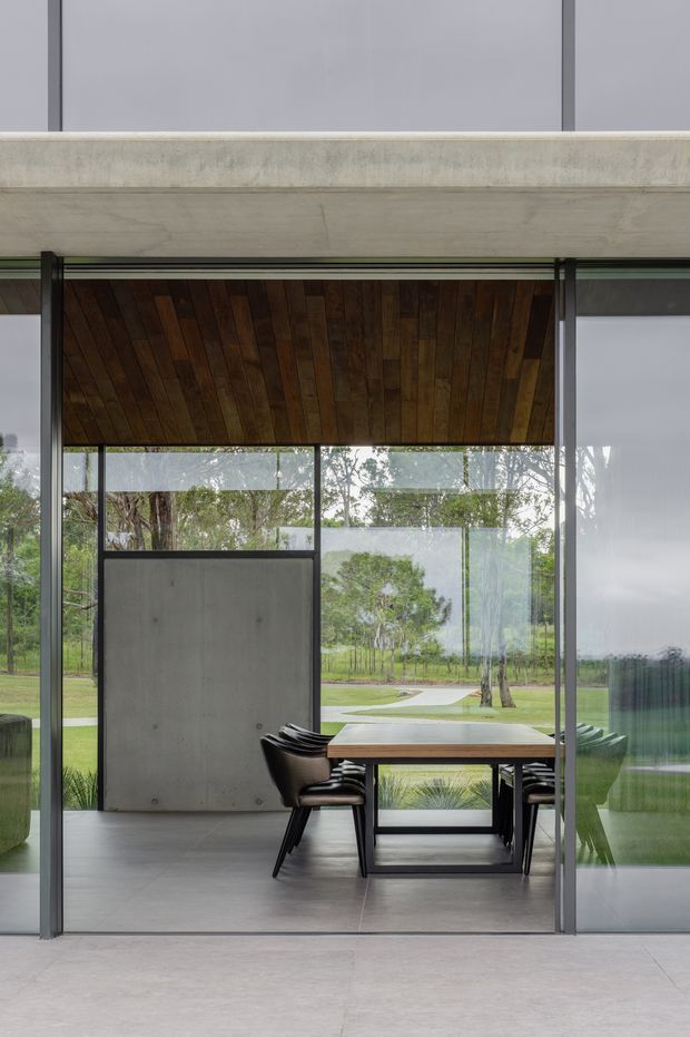 A refined contemporary dining area framed by full-height glass sliding doors. The space features a dark timber plank ceiling, a pale concrete wall panel, smooth grey flooring, and a substantial timber dining table with black sculptural chairs. The glazing opens views toward a landscaped garden, while slim dark metal framing creates a strong indoor-outdoor connection.