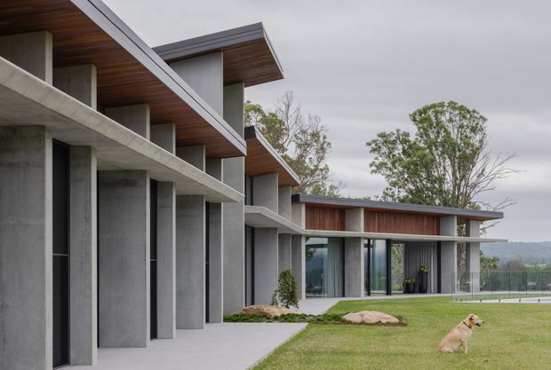 Exterior view of a contemporary multi-level residential building with strong horizontal roof planes, exposed concrete structural walls and columns, extensive floor-to-ceiling glazing, and warm timber soffits and cladding. The building sits beside a manicured lawn and landscaped grounds, with a covered entry walkway and distant landscape visible beyond.