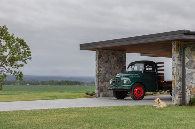 A rural property exterior with a low-profile flat-roof carport supported by rugged natural-stone columns and walls. The carport has a warm timber-lined soffit, dark metal roof edging, and horizontal timber screening at the rear. A vintage green truck with red wheels is parked beneath it, with expansive lawns and open farmland beyond under an overcast sky.