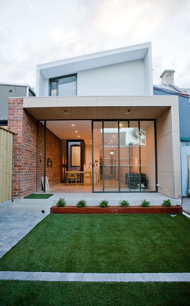 Rear exterior view of a compact contemporary urban home extension. The facade combines exposed red brick, pale smooth wall panels, black-framed floor-to-ceiling glazing, and a covered outdoor dining or entertaining area opening onto a small landscaped lawn. A simple projecting upper-level box and flat roof create a clean modern composition.