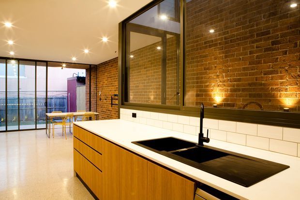 A warm, contemporary kitchen with a large timber island, white benchtop, black undermount sink and cooktop, white subway-tile splashback, and exposed brick walls. Black-framed internal glazing separates the kitchen from a dining area and balcony or terrace beyond. Recessed and under-cabinet lighting create a softly illuminated industrial-inspired interior.