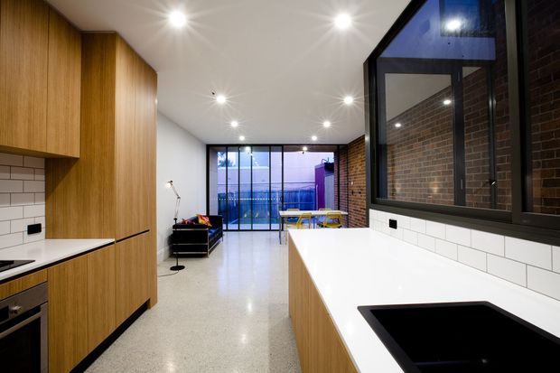 A contemporary open-plan kitchen opening into a living and dining area. The kitchen features warm timber cabinetry, white benchtops, white subway-tile splashbacks, a black-framed internal window, exposed brickwork, polished concrete or terrazzo-style flooring, and recessed ceiling lights. Glazing at the far end brings in daylight and provides views toward an exterior balcony or courtyard.
