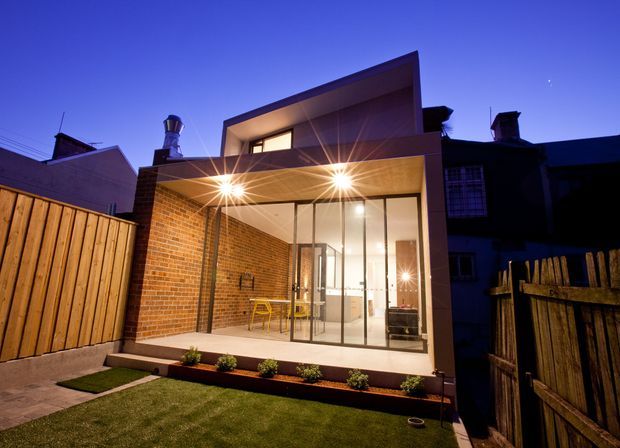 Twilight exterior view of a compact contemporary residential extension or rear dwelling. The design features a projecting upper-level box, a deep flat-roofed canopy, exposed red-brick walls, extensive floor-to-ceiling glazing with slim dark frames, and a small landscaped courtyard with timber fencing and artificial turf. Warm interior and exterior lighting contrasts with the deep blue evening sky.