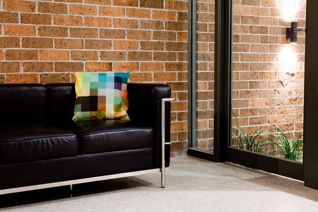 A modern interior corner featuring a black leather sofa with a colourful geometric cushion against an exposed reddish-brown brick wall. A tall black-framed glazed opening or internal window sits beside the sofa, with a wall-mounted light and planted area visible beyond the glazing. The room has a light grey carpet and a strong industrial character with warm accent lighting.