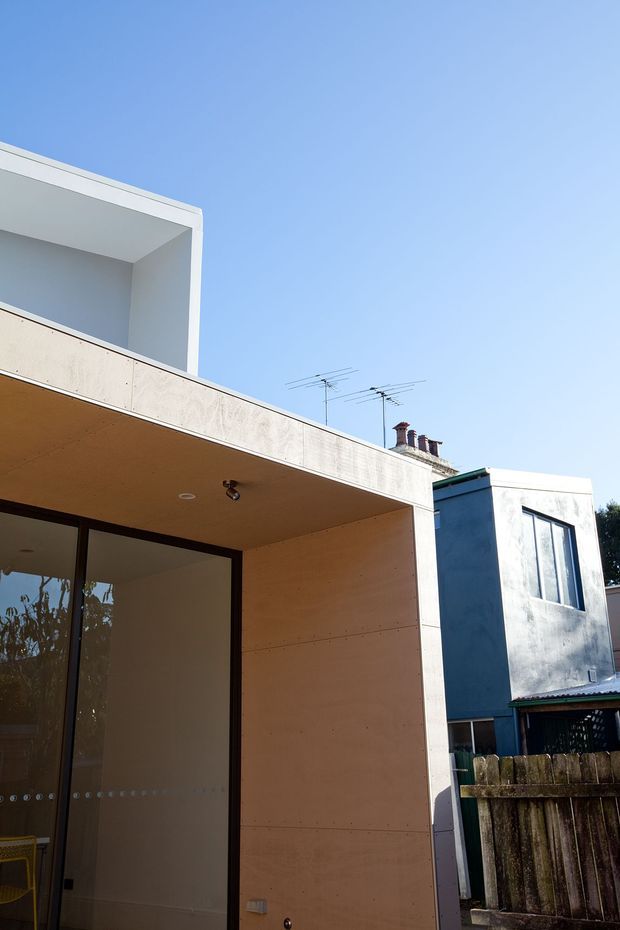 Exterior architectural view of a contemporary flat-roofed extension or dwelling, focused on a deep covered outdoor area with large glazed doors and pale timber-toned wall lining. A white rendered upper volume and neighbouring residential buildings are visible under a clear blue sky.