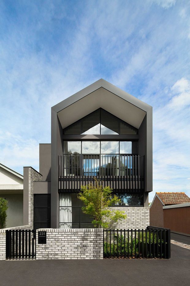 Front exterior view of a contemporary multi-storey urban dwelling with a prominent steep gabled roof form, dark rendered facade, extensive glazing, black vertical metal balustrades and a white brick boundary wall. The property includes a small front garden and gated entry, with neighbouring houses visible on either side.