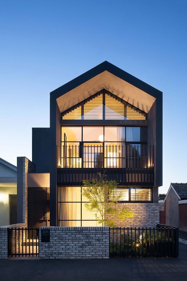 Front exterior view of a narrow multi-storey contemporary residence at dusk. The facade features a bold dark gabled frame, extensive floor-to-ceiling glazing, warm timber-toned gable lining, horizontal louvres, black metal balcony railings, and pale brickwork at the lower level and boundary wall. Warm interior lighting contrasts with the cool blue evening sky, while neighbouring houses are visible at the sides.